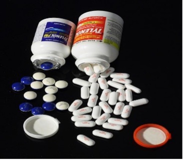 Two medicine bottles tipped over on a black surface with their caps off, spilling assorted tablets and capsules—white oblong pills, small white round tablets, and several blue-coated pills